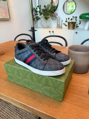 Gucci sneakers med monogram och ränder - Snygga Gucci sneakers i svart med GG-monogram över hela skon. Klassiska röda och blå ränder på sidan och metallic-detaljer i rött och blått på hälen. Skorna har vita sulor och svarta skosnören. Perfekta för dig som vill sticka ut med lyxig streetstyle. Box och kvitto ingår också!