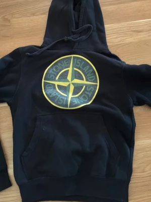 Svart hoodie från Stone Island - Säljer en svart hoodie från Stone Island med stor gul och grön logga på bröstet. Klassisk känguruficka framtill och justerbar huva med dragsko. Perfekt för dig som gillar streetwear och vill sticka ut med en ikonisk design.
