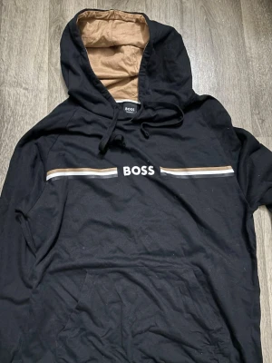 Svart hoodie från BOSS med beige detaljer - Svart hoodie från BOSS med beige och vita ränder över bröstet och BOSS-logga i vitt. Hoodien har en beige insida i huvan och dragsko. Känguruficka framtill och mjukt material som känns skönt mot huden. Perfekt för en avslappnad och stilren look.