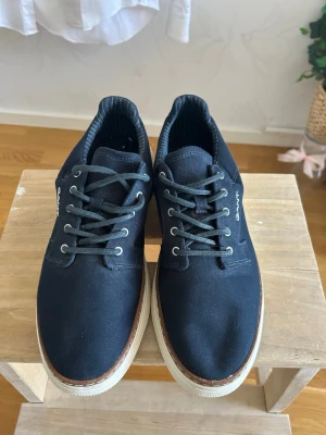 Marinblå Gant sneakers  - Säljer ett par Gant sneakers i marinblått. Skorna har vita sulor och snörning framtill. Gant-logotypen finns diskret på sidan av skorna. De är i storlek 44 och är i helt nyttskick. Skriv gärna om du vill ha fler bilder eller har några frågor, priset går såklart att diskutera.