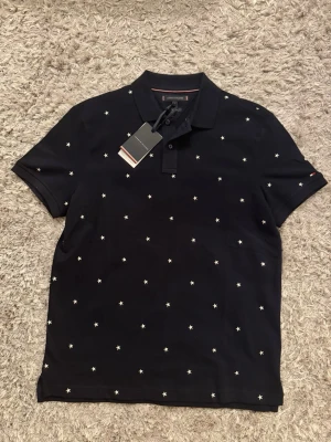 Svart piké med stjärnor Tommy Hilfiger - Svart pikétröja från Tommy Hilfiger med små vita stjärnor över hela plagget. Klassisk krage och korta ärmar. Tillverkad i ekologisk bomull för en skön och hållbar känsla. Perfekt för en stilren och avslappnad look. Helt nytt, storlek L ( 78-86kg ) 