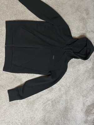 Svart hoodie från BOSS med dragkedja - Svart hoodie från BOSS med hel dragkedja framtill och diskret BOSS-logga på bröstet. Hoodien har huva med dragsko och två snedställda fickor. Snygg och stilren design som passar perfekt till streetwear eller chill. Är ej använd, helt ny 