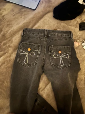 Svarta jeans med rosettbrodyr - Säljer ett par svarta jeans med raka ben och snygga broderade rosetter på bakfickorna. Jeansen har klassisk femficksmodell och kopparfärgade knappar. Perfekta för dig som vill ha en unik detalj på dina jeans. Low waist