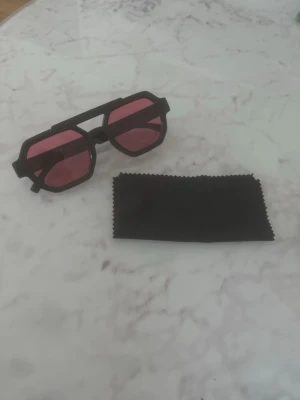 Svarta solglasögon med rosa glas - Säljer ett par trendiga solglasögon med svart, kantig båge och rosa glas. Glasögonen har en unik geometrisk form som sticker ut och passar perfekt för dig som vill ha en cool accessoar. Rengöringsduk medföljer. helt oanvända 