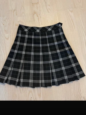 Svartvit rutig plisserad kortkjol - Snygg svartvit rutig kortkjol med plisserad design. Kjolen har ett klassiskt rutmönster och stängs med knapp i midjan. Perfekt för dig som gillar en preppy och trendig stil.