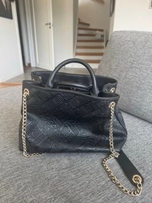 Svart handväska från Love Moschino - Snygg svart handväska från Love Moschino i präglat konstläder med textdetaljer. Väskan har guldfärgade kedjehandtag och en röd insida med innerfack. Perfekt storlek för det viktigaste och ger en cool vibe till din outfit.