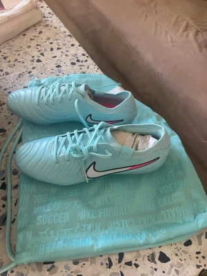 Nike turkosa fotbollsskor - Säljer ett par snygga turkosa fotbollsskor från Nike med vita och svarta detaljer samt en rosa linje på sidan. Skorna har klassisk snörning och är tillverkade i syntetmaterial med textil på insidan. Perfekta för dig som vill sticka ut på planen!