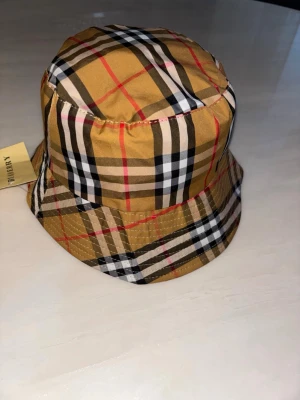 Burberry bucket hat i klassiskt rutmönster - Säljer en snygg bucket hat från Burberry med det ikoniska beige, svarta, vita och röda rutmönstret. Hatten är tillverkad i ett glansigt tyg och har en bred brätte. Insidan är svart med Burberry-logga runt kanten. Perfekt accessoar för att lyfta din outfit.