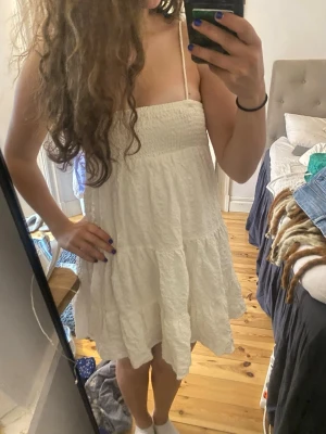 Vit sommarklänning  - Söt vit klänning med smala axelband och smockad överdel. Kjolen är volangig och har en luftig, avslappnad passform. Perfekt för varma dagar och enkel att styla med sneakers eller sandaler. Materialet är lätt och krinklat för en chill vibe.
