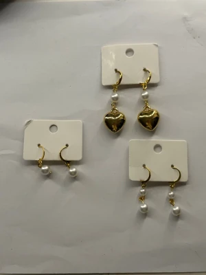 Guldörhängen med pärlor och hjärtan - Tre par örhängen i guld. Ett par med vita pärlor och hängande hjärtan, ett par med dubbla pärlor och ett par med enkel pärldetalj. Snygga accessoarer som ger en elegant touch till din stil. Perfekta för dig som gillar trendiga smycken med gulddetaljer.