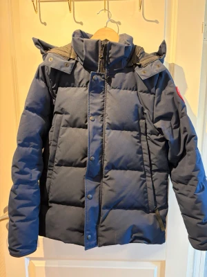 Mörkblå dunjacka från Canada Goose - Snygg mörkblå dunjacka från Canada Goose med hög krage, avtagbar huva och dragkedja med tryckknappar. Jackan har ett klassiskt quiltat mönster och logotyp på ärmen. Perfekt för kalla vinterdagar och riktigt varm tack vare dunfyllningen.