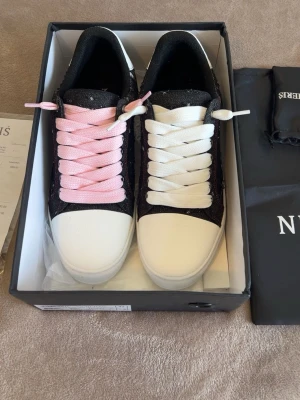 Svarta sneakers med rosa och vita snören - Unika sneakers med svart tyg och vita tåpartier. Ena skon har rosa snören och den andra vita, vilket ger en cool och personlig look. Yttermaterialet är en mix av textil och läderimitation. Perfekta för dig som vill sticka ut i mängden.