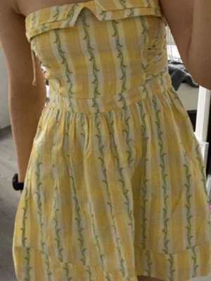 Gulvit axelbandslös sommarklänning - Söt axelbandslös kortklänning i gul och vit med blommigt mönster. Klänningen har en volangdetalj upptill och markerad midja. Perfekt för varma dagar och har en lätt, luftig känsla.🥰