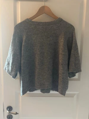 Grå stickad tröja med halvärm - Mysig grå stickad tröja med avslappnad passform och halvärmar. Tröjan har rund halsringning och är tillverkad i ett mjukt, melerat garn som ger en casual look. Perfekt att slänga på sig över en t-shirt eller skjorta.