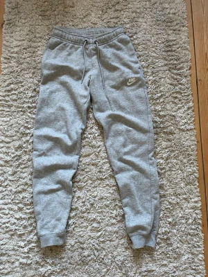 Gråa mjukisbyxor från Nike - Snygga gråa mjukisbyxor från Nike med klassisk logga på vänster ben. Byxorna har dragsko i midjan och muddar vid bensluten för en skön passform. Perfekta för chill eller träning, tillverkade i mjukt sweatshirtmaterial.
