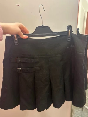 Svart kortkjol med spännen - Säljer en svart kortkjol från Bershka med två dekorativa spännen på sidan och veckad design. Kjolen har en klassisk och stilren look som passar till många olika outfits. Perfekt för dig som gillar en edgy men enkel stil. Lite kortare än vad som syns på bild .