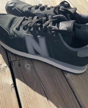 Svarta New Balance sneakers - Svarta sneakers från New Balance med grå detaljer och stor N-logga på sidan. Skorna har klassisk låg modell, svarta skosnören och vit sula med grå inslag. Ovandelen är i syntetmaterial och textil för en sportig look.