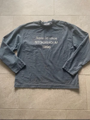 Acne Studios longsleeve - Snygg grå långärmad tröja från Acne Studios med tryck 'STOCKHOLM 1996' på bröstet. Tröjan har en avslappnad passform och ribbad rund halsringning. Perfekt för en casual och trendig look.