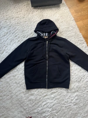 Svart hoodie från Burberry med dragkedja - Snygg svart hoodie från Burberry med dragkedja framtill och klassiskt rutigt foder i huvan. Hoodien har två fickor fram och diskret Burberry-logga på bröstet. Tillverkad i mjukt bomullsmaterial och har justerbar dragsko i huvan.