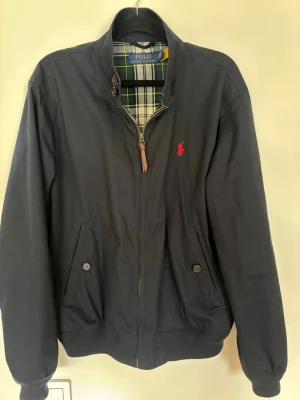 Polo Ralph Lauren Twill Jacket Marinblå  - Klassisk marinblå bomberjacka från Polo Ralph Lauren med röd broderad logga på bröstet. Jackan har dragkedja, två framfickor med knappar och rutigt foder i grönt, vitt och gult. Ribbad mudd vid ärmslut och nederkant. Perfekt för vår och höst.