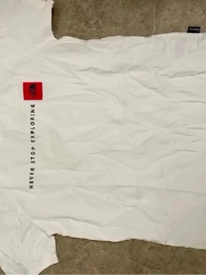 Vit t-shirt från The North Face - Vit t-shirt från The North Face med svart text 'Never Stop Exploring' längs sidan och en röd logga på bröstet. Klassisk passform och rund hals. Perfekt för en avslappnad och stilren look.