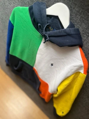 Färgglad hoodie från Polo Ralph Lauren - Säljer en riktigt snygg hoodie från Polo Ralph Lauren i blockfärger: grön, blå, vit, orange, gul och marinblå. Klassisk huva med dragsko, magficka och broderad logga på bröstet och ärmen. Perfekt för dig som vill sticka ut med en unik och färgstark stil.