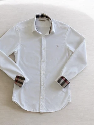 Vit skjorta från Burberry med rutiga detaljer - Vit skjorta från Burberry i bomull med klassiska rutiga detaljer på insidan av krage och manschetter. Skjortan har lång ärm, knappar framtill och en liten broderad logga på bröstet. Perfekt för en stilren look.