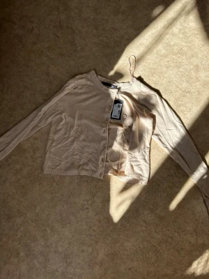 Beige cropped kofta från laget 157 - Snygg beige cropped kofta från H&M med rund halsringning och långa ärmar. Koftan har knappar framtill och ribbade muddar vid ärmslut och nederkant. Perfekt att slänga över en topp för en chill look.