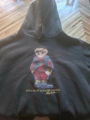 Svart hoodie Polo Bear Ralph Lauren - Svart hoodie från Ralph Lauren med ikoniska Polo Bear-trycket framtill. Klassisk huva och avslappnad passform, perfekt för en chill och stilren look. Säljer denna för 850kr men pris kan diskuteras. Nypris: 2995kr