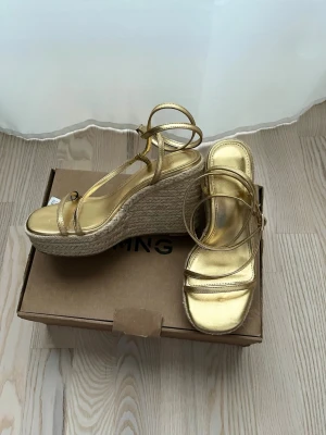 Guldiga wedge-pumps med snörning, storlek 35, 10cm, Mango - Snygga guldiga pumps från Mango med hög kilklack i flätad jute och tunna remmar som knyts runt vristen. Skorna har öppen tå och metallic-finish, perfekta för att sticka ut. Klackhöjd 10 cm. 