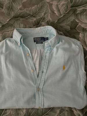 Ljusblå skjorta från Polo Ralph Lauren - Klassisk ljusblå skjorta från Polo Ralph Lauren med button-down krage och broderad gul logga på bröstet. Skjortan har långa ärmar och knäppning framtill. Perfekt för en stilren och avslappnad look.