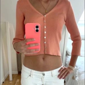 Persikofärgad ribbad topp - Trendig persikofärgad ribbad kofta med v-ringning och dekorativa pärlemorknappar framtill. Koftan har trekvartsärmar och croppad passform som ger en modern look. Perfekt att styla med höga jeans eller kjol. Ganska tunn i materialet 💓