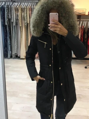Svart parkas med äkta päls från hollies - Snygg svart parkas från hollies med stor huva kantad av beige päls. Jackan har guldfärgade dragkedjor och knappar, två stora fickor framtill och en bröstficka med dragkedja. Figurnära passform och dragsko i midjan. Perfekt för kalla dagar. Använd 1 vinter 