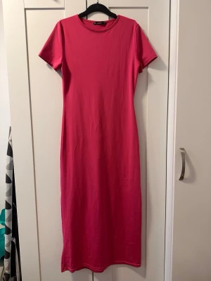Rosa långklänning - En enkel och stilren rosa långklänning. Klänningen har rund halsringning och korta ärmar. Tillverkad i mjuk bomull som ger en bekväm känsla. Perfekt för dig som gillar minimalistisk och avslappnad stil.