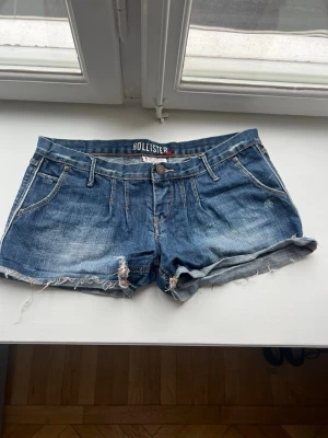 20s jeans shorts Hollister - Säljer ett par klassiska blå jeansshorts från Hollister med råa kanter och femficksmodell. Shortsen har låg midja och slitna detaljer för en avslappnad look. Perfekta till sommaren och lätta att matcha med allt.