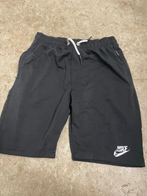 Svarta shorts från Nike med snörning - Säljer ett par svarta shorts från Nike med vit snörning i midjan och logga på ena benet. Shortsen har en avslappnad passform och är perfekta för träning eller chill. Tillverkade i mjuk bomull för skön känsla.