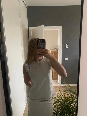 Vit spetsblus med ärmlös design - Söt vit blus i spets med ärmlös design och fina spetsdetaljer vid axlarna. Blusen har rund halsringning och ett romantiskt uttryck, perfekt för dig som gillar en feminin stil.