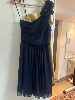 Marinblå oneshoulder klänning - Snygg marinblå oneshoulder klänning med tunna axelband och en stor dekorativ blomma på axeln. Klänningen har draperad överdel och ett flytande, lätt tyg i kjolen. Perfekt för speciella tillfällen när du vill sticka ut.