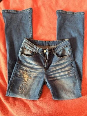 Blå jeans med fjärilsbrodyr - Snygga blå jeans med utsvängda ben. På ena benet finns en broderad fjäril med dekorativa detaljer och diamanter. Klassisk femficksmodell med knapp och dragkedja. 