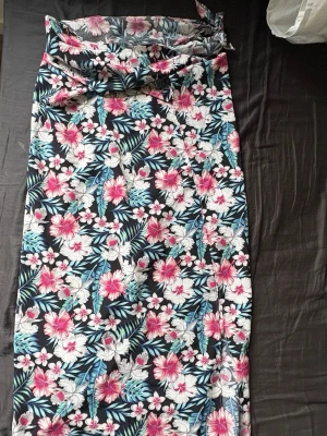 Blommig omlottkjol med knytband - Snygg långkjol med blommigt mönster i rosa, vitt och blått på svart botten. Kjolen har omlottmodell och knytband i midjan. Man kan säkert använda den som topp om man vet hur man ska knyta. Perfekt som strand cover up. 