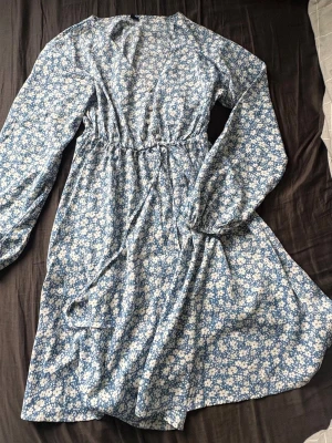 Blommig blå kortklänning med knyt - Söt blå kortklänning med vitt blommönster, v-ringning och pärlknappar framtill. Klänningen har långa ärmar och ett knytband i midjan som dekoration. Perfekt för sommar!