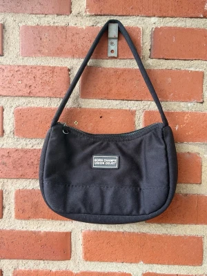 Svart axelväska - En svart handväska från Born Champs union odjet med enkel design och ett litet vitt logotypmärke framtill. Väskan har en mjuk, rundad form och är tillverkad i tygmaterial. Dragkedja upptill och en smal axelrem gör den smidig att bära.
