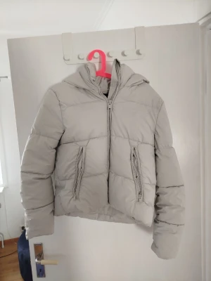 Beige pufferjacka från Zara - Snygg vit pufferjacka från Zara med stor huva och två framfickor med dragkedja. Jackan har en rak, boxig passform och stängs med dragkedja framtill. Perfekt för kalla vinterdagar och ger en trendig look.