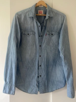 Ljusblå jeansskjorta från Levi's - Säljer en klassisk ljusblå jeansskjorta från Levi's med två bröstfickor och knappar framtill. Skjortan har lång ärm och snygga detaljer vid axlarna. Perfekt för dig som gillar en avslappnad och tidlös look. Slim fit! 