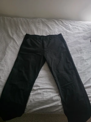 Svarta chinos med rak passform - Säljer ett par svarta chinos med rak passform och klassisk design. Byxorna har knappgylf och bälteshällor, samt en gul detalj på insidan av linningen. Materialet ser ut att vara bomull eller bomullsblandning.