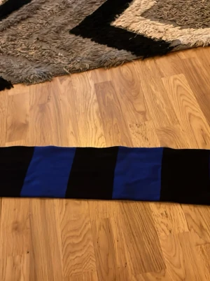 Sirius halsduk blå och svart - Säljer en snygg halsduk från Sirius med breda blå och svarta ränder. Halsduken har fransar i ändarna och Sirius klubbmärke broderat på båda sidor. Perfekt för dig som vill visa ditt stöd för laget och hålla dig varm på matchen.