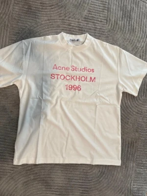 Vit topp från Acne Studios med tryck - Vit topp från Acne Studios med rosa texttryck 'Acne Studios STOCKHOLM 1996' på framsidan. Klassisk rund hals och korta ärmar. Tillverkad i mjuk bomull för en bekväm och stilren känsla.