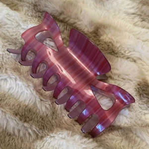 Stor Rosa Hårklämma i Plast - Säljer en stor hårklämma med snyggt randigt mönster i olika rosa nyanser. Klämman har en klassisk form med breda grepp och öppna detaljer på sidorna, perfekt för att hålla upp håret på ett trendigt sätt.