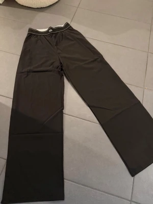 Svarta suit up byxor Alo S 36 - Säljer ett par svarta mjukisbyxor från Alo med bred resår i midjan och diskret logga framtill. Byxorna har raka, vida ben och är gjorda i ett mjukt och stretchigt material som känns riktigt skönt mot huden. Perfekta för chill eller träning.