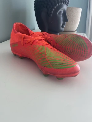 Puma orangea fotbollsskor - Säljer ett par grymma orangea fotbollsskor från Puma med gröna detaljer och dobbar. Skorna har snörning och är tillverkade i ett lätt syntetmaterial som ger bra bollkontroll och grepp på planen. Perfekta för dig som vill sticka ut på fotbollsplanen!